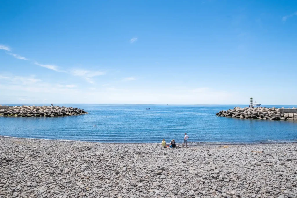 Rocky beach with calm blue waters and distant breakwater structures.